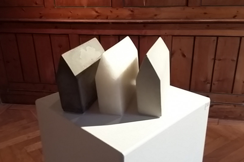 Skulptur: drei identisch geformte Spitzdach-Häuser aus drei unterschiedlich farbigen Materialien (dunkelgrau, weiß, hellgrau) 