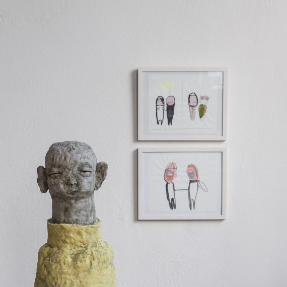 Tonfigur mit gelbem Pulli, dahinter zwei Skizzen mit Figuren an Wand