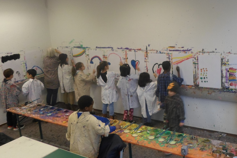 Kinder malen in Malerkitteln mit bunten Farben auf große weiße Blätter an der Wand