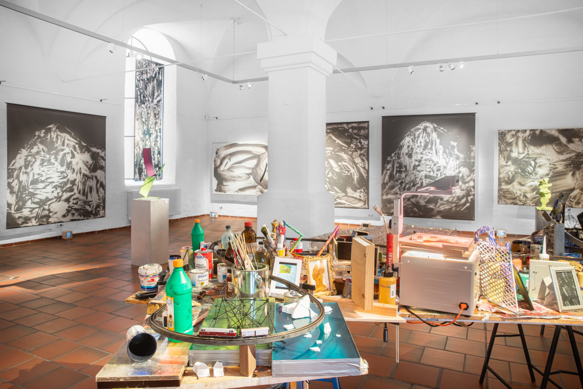 Installation in der Raummitte aus Ateliertischen - überfüllt mir Arbeitsmaterialien und weiteren inszenierten Gegenständen, große Schwarz-Weiß-Bilder an den Wänden