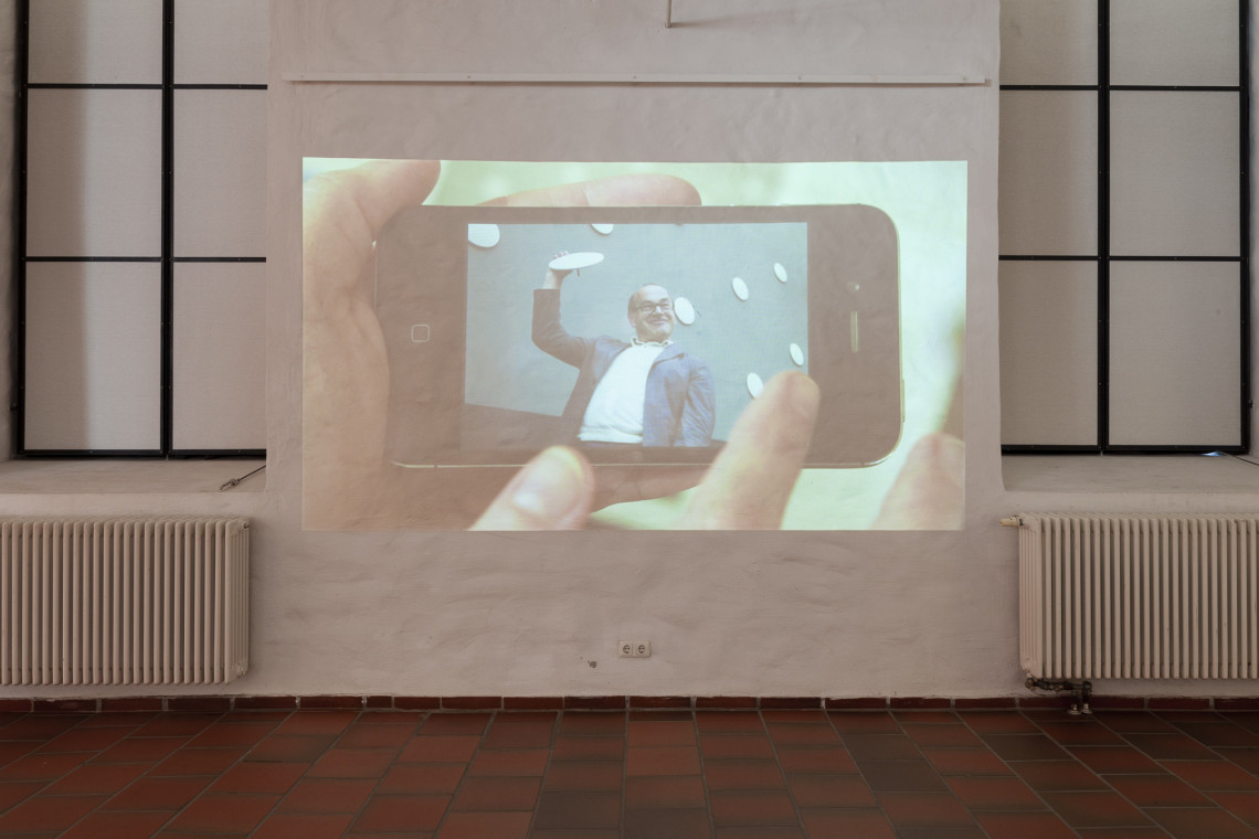 Videoprojektion: Hand hält Smartphone, auf Display ist ein Mann zu sehen, der stolz einen weißen Teller in die Höhe hält