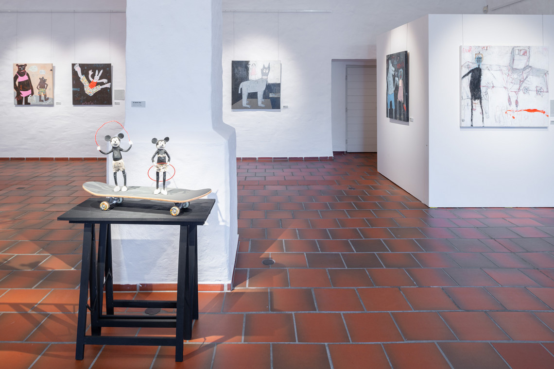 Blick in Ausstellungsraum, im Vordergrund eine Installation mit zwei Mickey Mäusen auf einem Skateboard, im Hintergrund Bilder an Stellwänden