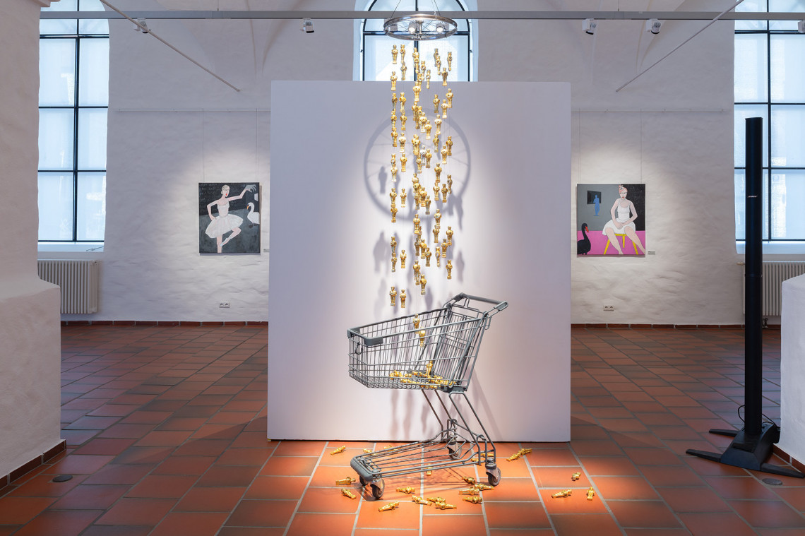 Blick in Ausstellungsraum mit einem leeren Einkaufswagen vor einer Stellwand und einem goldenen Mobile darüberhängend